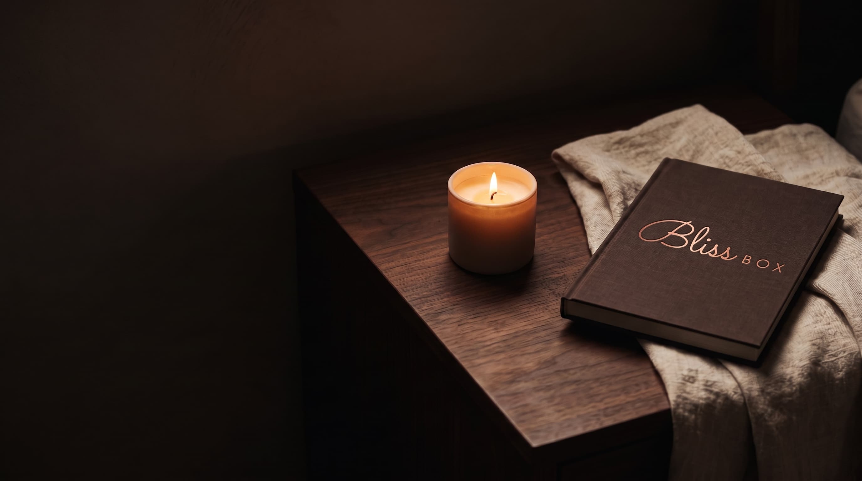 A lit candle and a cloth bound book on a dark walnut bedside table, beside a linen sheet