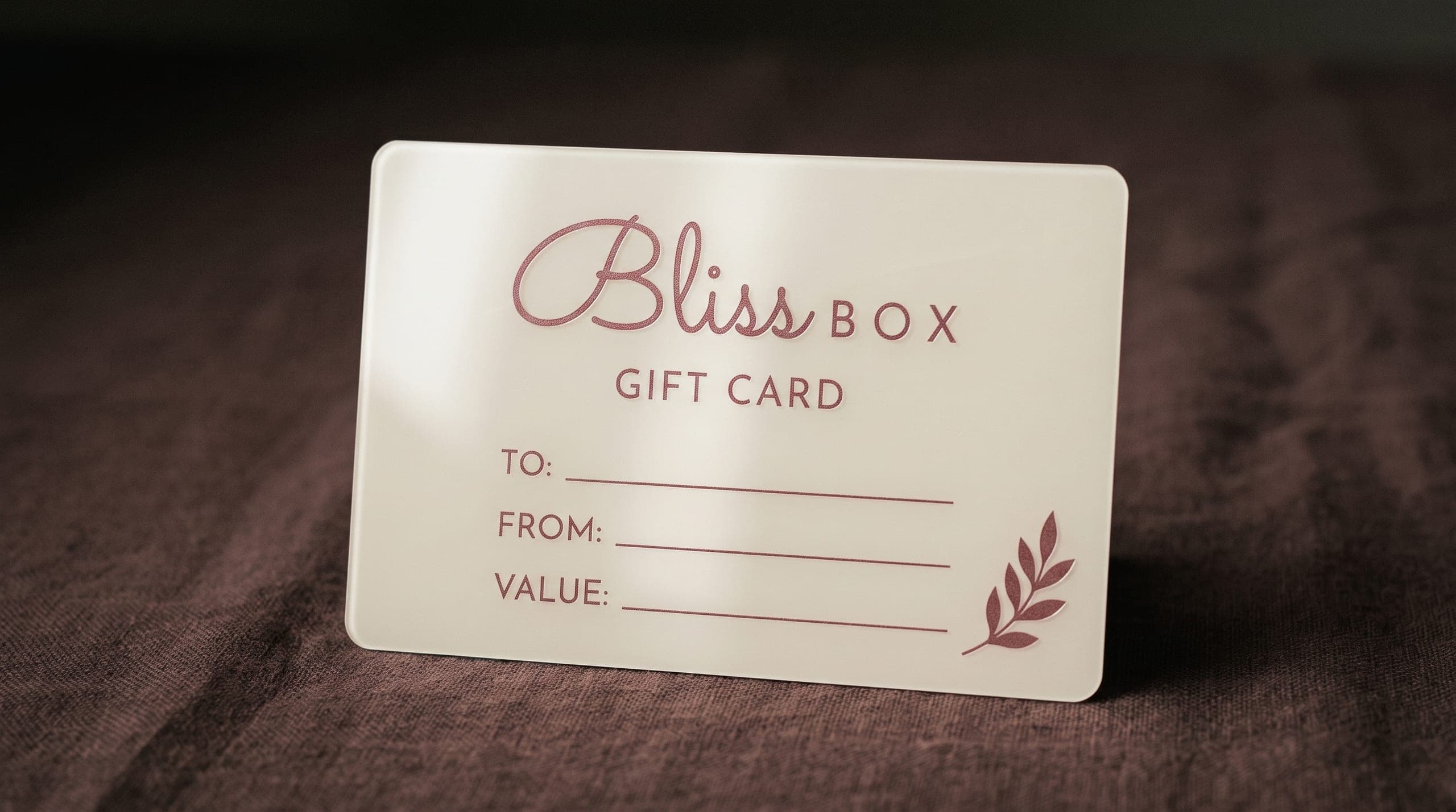 A Bliss Box gift card, printed with the wordmark and lines for to, from and value, standing on dark linen
